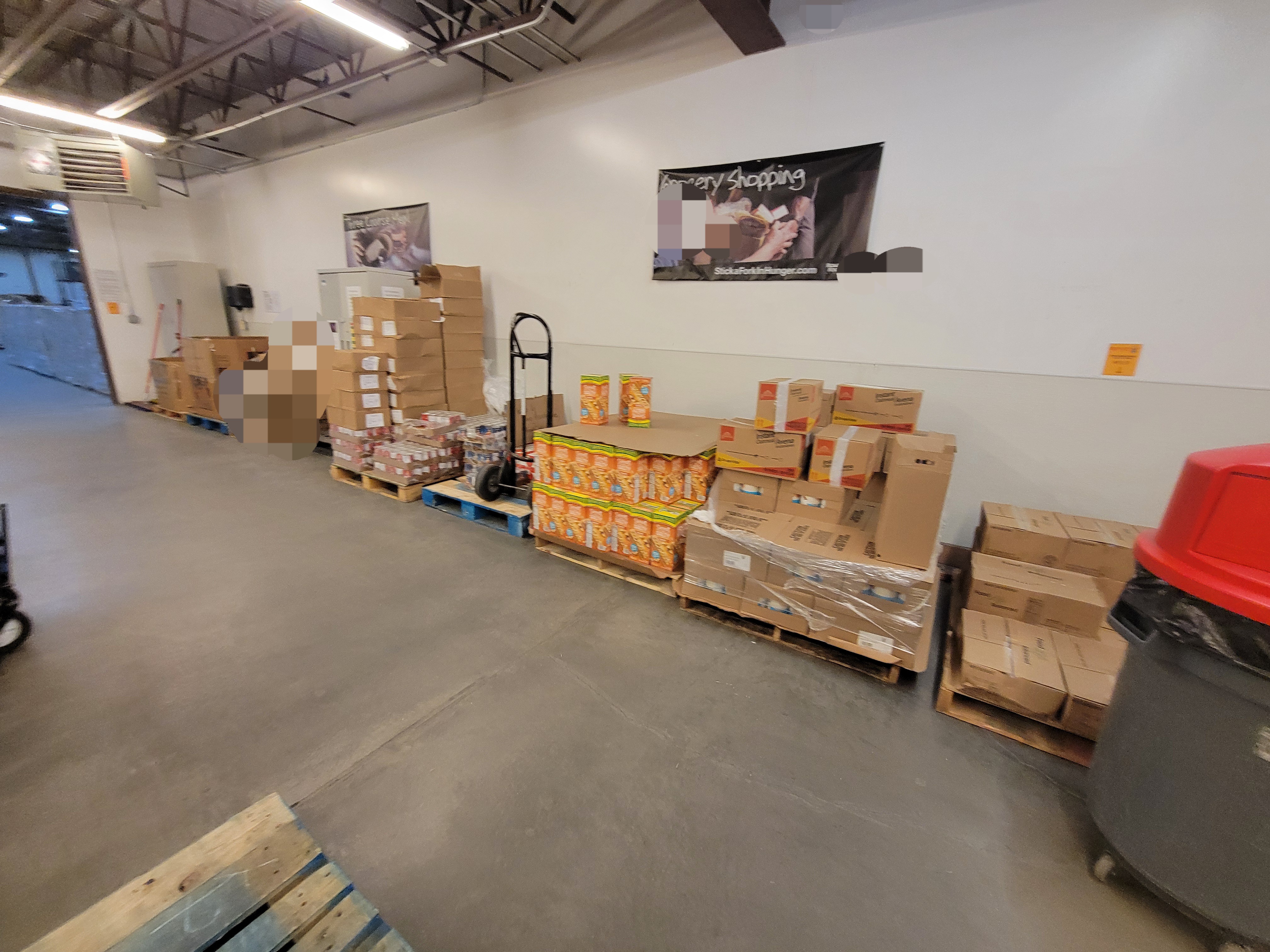 Pallets of food with a trash can near by, and hand truck on top of one.